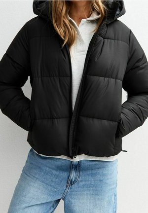 Doudoune noire à capuche zippée à mi-hauteur, portée sur un polo blanc avec un jean bleu clair, les mains dans les poches de la veste.