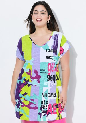 Kvinde med langt mørkt hår iført en farverig, abstrakt mønstret kortærmet kjole med blandede tekst- og grafikmønstre mod en hvid baggrund.