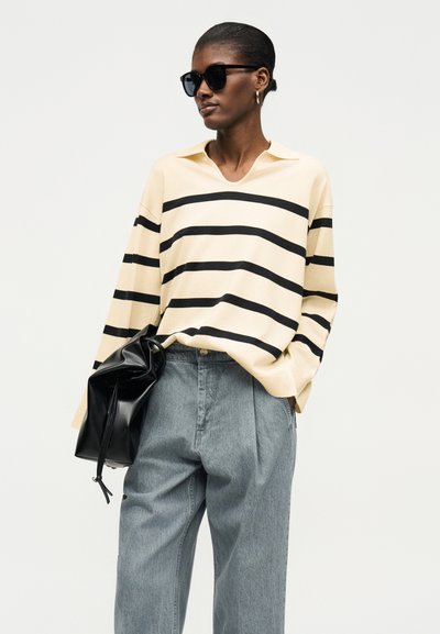 Γυναίκα με μαύρα γυαλιά ηλίου, κρεμ πουλόβερ με μαύρες ρίγες, φαρδιά μπλε τζιν, κρατώντας μια μαύρη δερμάτινη τσάντα, ποζάροντας σε λευκό φόντο.