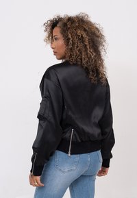 Zwarte satijnen bomberjack met een aansluitende onderrand, ritszakken en geribbelde mouwen; heeft een gladde textuur en een licht oversized silhouet.