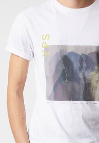 Bílá bavlněná tričko s ohrnutými rukávy, které má vybledlý grafický potisk; text "YPS" v světle žluté barvě a umělecké znázornění překrývajících se postav.