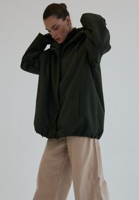 Przesadnie duża ciemnozielona kurtka z zamkiem błyskawicznym, elastycznym dołem i dwoma bocznymi kieszeniami. Nosi się ją na czarnym golfie, zestawiona z beżowymi spodniami.