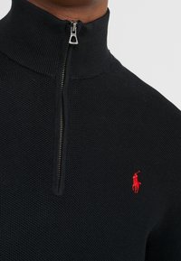 Svart zip-up tröja med hög krage, texturerat stickat material och en röd logotypaccent på vänster bröst. Har silverfärgad dragkedja.