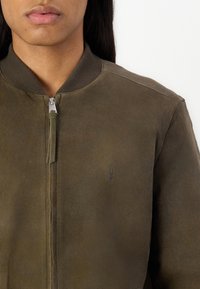 Giubbotto bomber in pelle verde oliva scuro con chiusura lampo frontale, colletto a coste e logo goffrato discreto sul petto. Texture liscia.