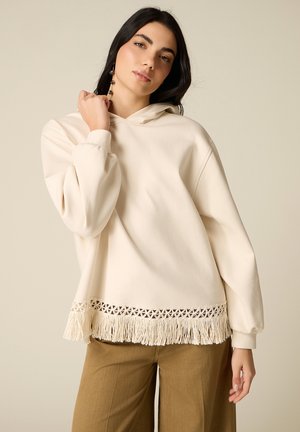 Donna con lunghi capelli scuri che indossa una felpa con cappuccio color crema con dettagli frangiati e pantaloni marroni, su uno sfondo beige uniforme.