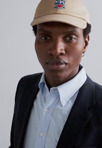 Beige kasket med logo, lyseblå skjorte med knapper, mørk blazer og ørestik. Nærbillede portræt, neutral baggrund.