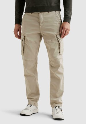 Man draagt beige cargobroek met zijzakken en witte sneakers, staand tegen een effen lichte achtergrond.