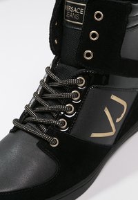 Baskets montantes noires en mélange de daim et de matériaux brillants, avec des accents dorés, un logo bien en vue et des lacets à motifs.