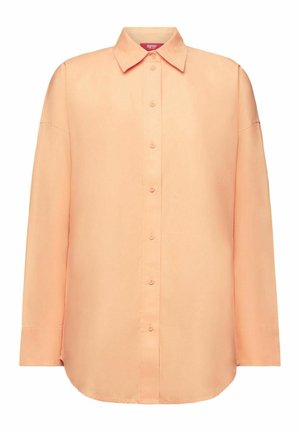 Personne portant une jupe rose, une chemise pêche, une longue veste en jean, une écharpe rose, des baskets beige et des chaussettes blanches avec des cheveux tressés visibles.