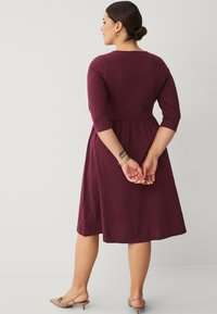 Bordeaux, knielange jurk met driekwart mouwen, een getailleerde bodice en een ontspannen rok. Gladde stof en ronde halslijn, gedragen met tan flats.