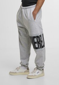 Sivé bavlnené tepláky s čiernou grafikou na dolnej nohe, s bočnými vreckami a elastickým pásom. Nosené s béžovými teniskami.