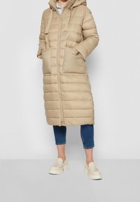 Beige prošívaný dlouhý kabát s kapucí, s předním zipem, dvěma velkými kapsami a horizontálním prošíváním. Nositý přes modré džíny.