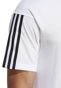 T-shirt de sport blanc en tissu texturé, avec des accents noirs rayés sur les manches et un design de col rond simple.