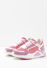 Růžové atletické tenisky z kombinace semiše a textilních materiálů. Mají bílé tkaničky, bílé logo a robustní strukturovanou podrážku.