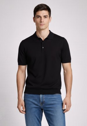 Młody mężczyzna w czarnej, krótkim rękawem koszulce polo i niebieskich dżinsach, stojący na tle jednolitego jasnego tła.