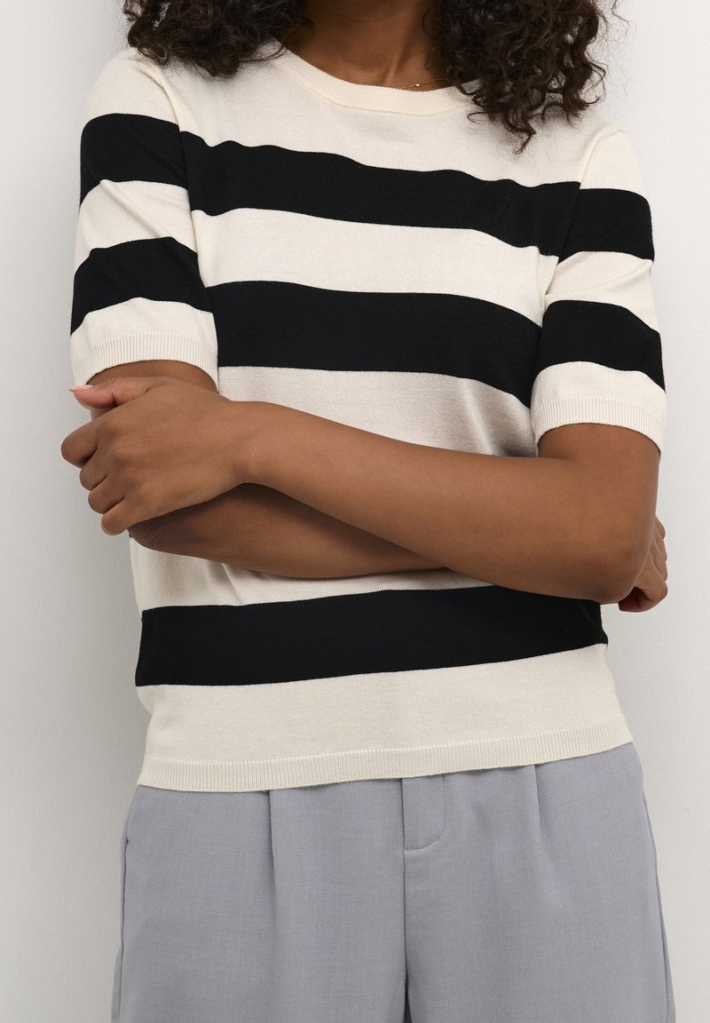 Personne portant une chemise à rayures horizontales noires et blanches à manches courtes et un pantalon gris clair, les bras croisés devant un mur blanc.