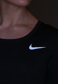 Musta, tiukasti istuva urheilupaita, jossa on teksturoitu pinta ja vasemmassa olkapäässä näkyvä suuri valkoinen Nike-logo.