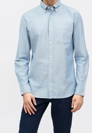 Mann trägt ein hellblaues Hemd mit Knöpfen und Brusttasche sowie dunkle Hose, steht vor einem schlichten weißen Hintergrund.