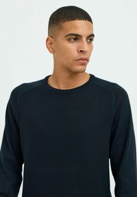 Pull-over long bleu foncé à manches longues avec une texture côtelée et un col rond. Présente un tissu lisse contrastant sur les épaules.