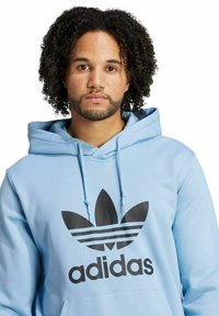 Felpa leggera blu chiaro con un logo trefoil nero di Adidas e testo. Presenta un cappuccio con cordini e una tasca frontale a marsupio, realizzata in tessuto morbido.