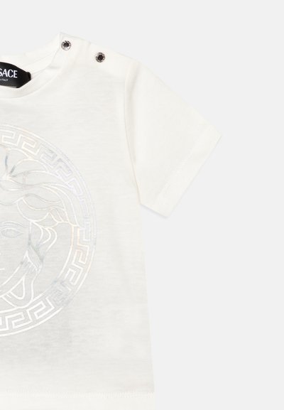 Camiseta blanca de manga corta con un emblema brillante de Medusa y un patrón de clave griega. Detalles con botones a presión en los hombros. Material suave de algodón.