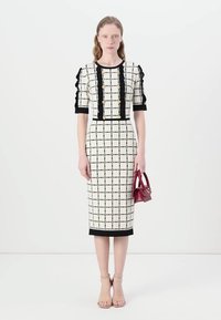 Rochie albă cu un model de plasă neagră și nasturi aurii, design ajustat, cu mâneci scurte volănașe și tiv negru. Modelul ține în mână o poșetă roșie mică.