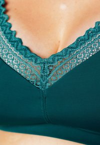 Reggiseno color teal con bordo in pizzo a onda lungo il décolleté, tessuto liscio con una leggera lucentezza e un classico design a forma di V.