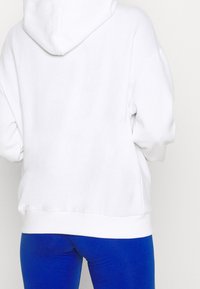 Sweat à capuche blanc en tissu de coton doux avec une poche kangourou et une capuche à cordon de serrage ajustable. Associé à des leggings bleus.