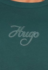 Maglione verde a coste con logo "Hugo" ricamato in argento sul petto. Il materiale sembra morbido e strutturato.