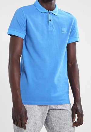 Mann in einem leuchtend blauen Poloshirt mit kurzen Ärmeln und schwarzen Knöpfen, kombiniert mit hellgrauen, strukturierten Hosen vor einem schlichten Hintergrund.