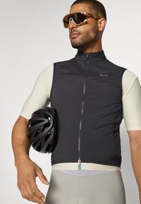 Gilet sans manches noir avec un col haut, fermeture éclair et logo. Chemise ajustée de couleur claire en dessous. Casque de vélo noir dans le bras du modèle.