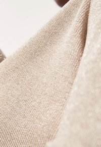 Tessuto a maglia beige con una leggera lucentezza, caratterizzato da una trama fine e una caduta morbida. La superficie presenta delicate filature brillanti in tutta la sua estensione.