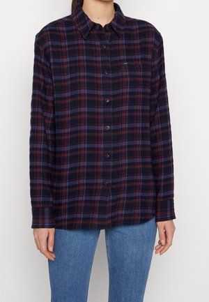 Woman wearing dark blue and burgundy plaid button-up shirt with chest pocket, paired with light blue jeans, standing against neutral background.