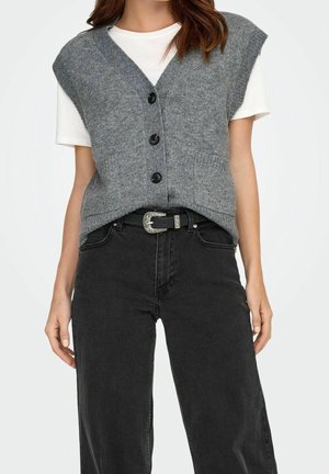 Femme portant un gilet en tricot gris boutonné sur une chemise blanche à manches courtes, associé à un jean noir taille haute et une ceinture noire avec une boucle argentée.