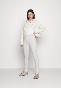 Casaco cropped bege claro sobre uma blusa branca, combinado com jeans cinza claro de cintura alta e sandálias em tom bege com detalhe de laço. Acessório simples.