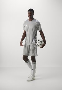Šedý futbalový dres Adidas s krátkymi rukávmi, pruhovanými akcentmi a zodpovedajúcimi šortkami. Hráč drží čierno-biely futbal.