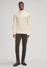 Kremowy sweter z golfem i teksturowanym ribbingiem, w połączeniu z dopasowanymi oliwkowymi spodniami i brązowymi botkami za kostkę, stojący na neutralnym tle.