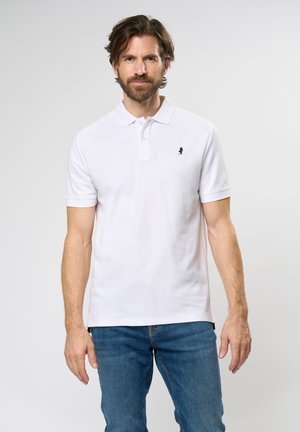 Weißes Polo-Shirt mit kurzen Ärmeln, traditionellem Kragen und einem kleinen Marineblauen Logo auf der Brust. Getragen mit blauen Jeans.