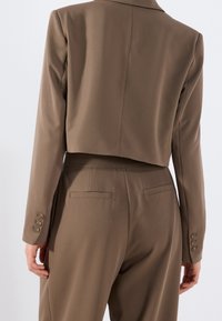Brązowa krótka marynarka w zestawie z dopasowanymi spodniami. Charakteryzuje się gładką tkaniną, dopasowanym krojem oraz guzikami na mankietach i tylnych kieszeniach spodni.