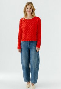 Roter Strickpullover mit Zopfmuster und langen Ärmeln, kombiniert mit locker sitzenden blauen Jeans und hellen Pumps. Der Pullover ist mit texturierten Wellenmustern versehen.