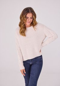 Hellrosa gestrickter Pullover mit geripptem Saum und Zopfstrickdetails an den Ärmeln, kombiniert mit dunkelblauen, engen Jeans. Weiche Textur.
