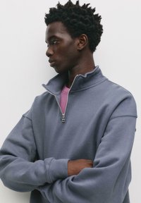 Felpa a zip blu scuro con colletto alto e texture morbida. Presenta maniche lunghe e larghe e una zip in metallo argentato. Indossata sopra una maglietta rosa.