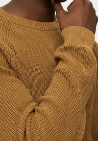 Hnědý pletený svetr s kulatým výstřihem, texturovanou tkaninou a žebrovanými manžetami, který se vyznačuje pohodlným střihem a podrobným prošíváním.