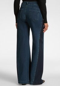 Vaqueros de pierna ancha en azul oscuro, con paneles laterales en contraste, costuras decorativas y dos bolsillos traseros. Textura suave, diseño casual.