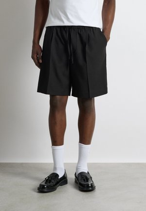 Homme portant un short noir à cordon, des chaussettes blanches et des mocassins noirs à pampilles, debout devant un fond uni.