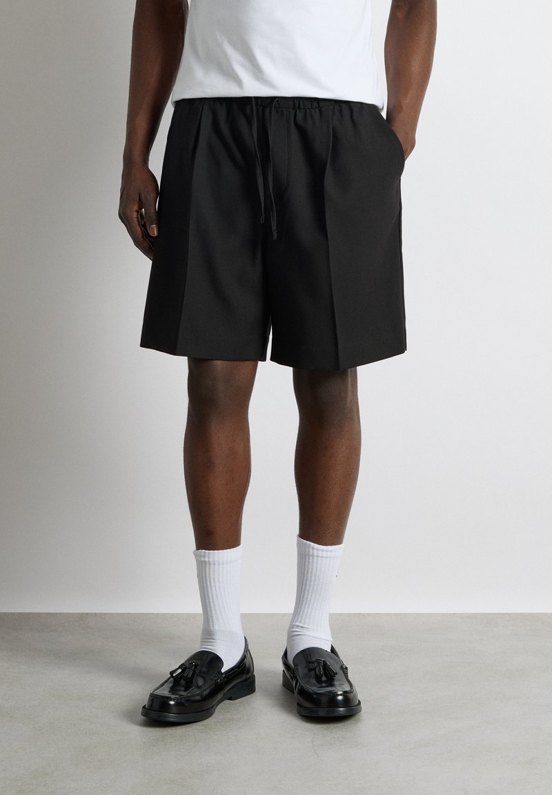 Homme portant un short noir à cordon, des chaussettes blanches et des mocassins noirs à pampilles, debout devant un fond uni.