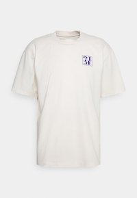 T-shirt crème à manches courtes en coton doux avec une coupe décontractée, arborant un logo géométrique violet sur la poitrine gauche. Design simple.