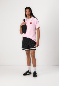 Pink short-sleeve t-shirt with logo, black patterned shorts, black shoulder bag, and black sneakers with white stripes. Casual athletic style.
