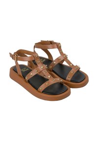 Bruine synthetische sandalen met meerdere banden, voorzien van gouden studs, een platte zwarte zool en een gespsluiting rond de enkel.