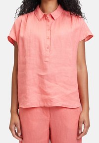 Nainen, jolla on päällään korallinvärinen, napitettava, lyhythihainen pellavapaita ja vastaavat pellavahousut, valkoista taustaa vasten.